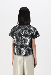Maglietta nera a maniche corte con motivo astratto metallizzato argento, caratterizzata da una vestibilità rilassata e dettaglio a pieghe sul retro, abbinata a pantaloni chiari.