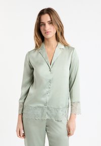 Zielona satynowa góra od piżamy z wyciętym kołnierzem, zapięciem na guziki z przodu oraz koronkową lamówką na rękawach i dole, o gładkiej teksturze.
