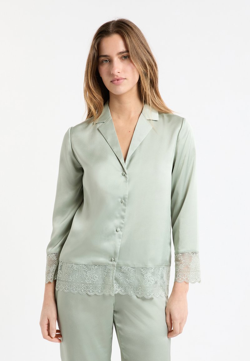 Zielona satynowa góra od piżamy z wyciętym kołnierzem, zapięciem na guziki z przodu oraz koronkową lamówką na rękawach i dole, o gładkiej teksturze.
