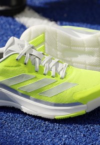 Bright yellow and white Adidas athletic shoe with white laces on textured blue surface.