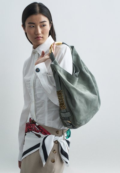 Ázijská žena v bielej košeli a béžovej sukni s viazanou šálou, nesúca veľkú šedú tašku Moschino cez rameno, pozerajúca sa bokom.