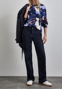 Blusa a fantasia floreale nei colori blu navy, crema e borgogna; abbinata a jeans a gamba larga blu scuro e sneakers bianche. Tessuto leggero, vestibilità casual.