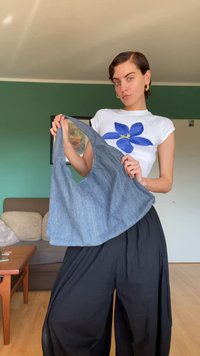 Kerek formájú farmer táska, egyetlen pánttal, egy személy kezében bemutatva. Világos kék-szürke színű pamut anyag.