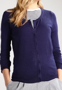 Cárdigan azul marino hecho de tela de punto, con mangas largas, cuello redondo y cierre de botones en la parte delantera. Camiseta a rayas debajo.
