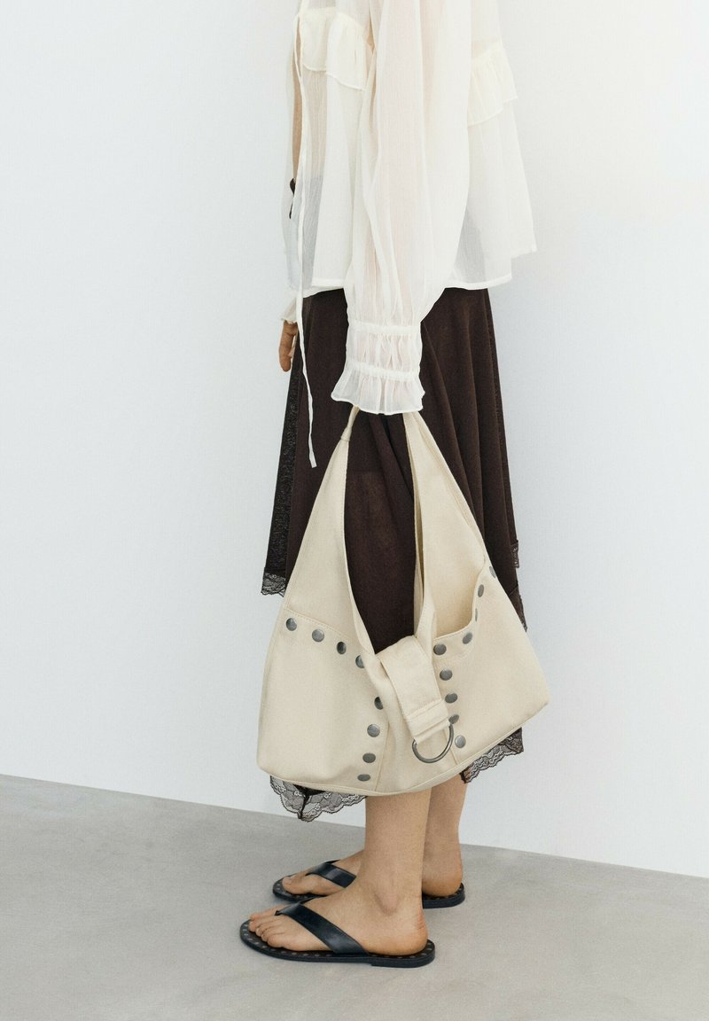 Personne portant un chemisier blanc et une jupe foncée, tenant un sac à main beige clouté, debout sur un sol gris avec des sandales slides noires.