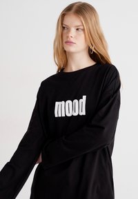 Crna košulja dugih rukava izrađena od mekanog materijala, s velikim bijelim grafičkim natpisom "mood" na prednjoj strani. Opušteni kroj s spuštenim ramenima.