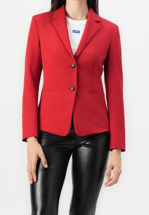 Vrouw draagt een nauwsluitend rode blazer met twee knopen over een witte top, gecombineerd met zwarte leren broek tegen een effen witte achtergrond.