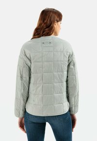 Hellegraue gesteppte Jacke mit boxy Passform, langen Ärmeln und Rundhalsausschnitt. Besitzt eine glatte Oberfläche und zwei Druckknopfdetails auf der Rückseite.