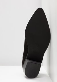 Schwarzer Wildleder-Schuh mit hohem Absatz und spitzem Zehenbereich, glattem Absatz und strukturiertem Außensohle, vor einem weißen Hintergrund platziert.