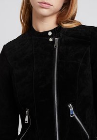 Svart mockajacka med ståkrage, silverfärgad dragkedja och sidfickor med dragkedja samt texturerade axeldetaljer.