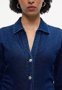 Τζιν πουκάμισο σε σκούρο μπλε με κολάρο, μπροστινό κλείσιμο με κουμπιά και διακριτές ραφές. Λείο ύφασμα και εφαρμοστό κόψιμο.