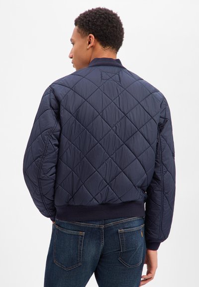 Mann, der von hinten zu sehen ist, trägt eine marineblaue gesteppte Bomberjacke und dunkelblaue Jeans vor einem schlichten weißen Hintergrund.