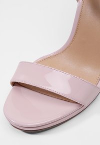 Sandal con tacco alto in vernice rosa con una larga fascia sulla parte frontale e una finitura liscia e lucida. La suola è di un beige chiaro e texturizzata.