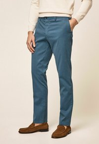 Teal cotton trousers with a slim fit, featuring side pockets, a zip fly, and a button closure. Worn with brown suede loafers.