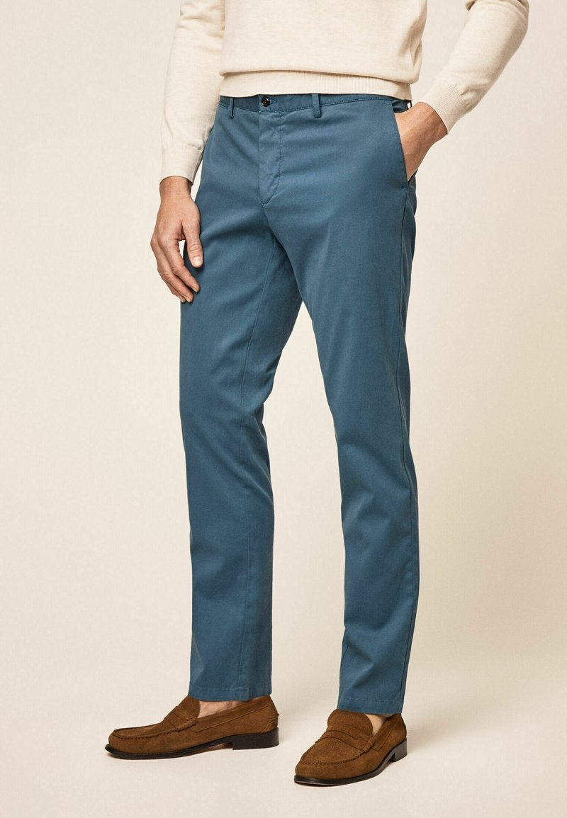 Teal cotton trousers with a slim fit, featuring side pockets, a zip fly, and a button closure. Worn with brown suede loafers.