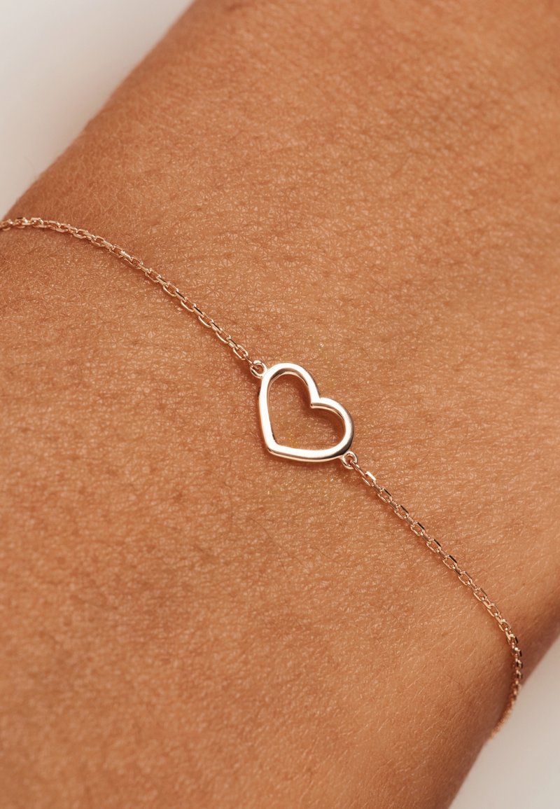 Roze gouden kettingarmband met een open hartbedel. De armband heeft een delicate, dunne uitstraling en wordt gedragen om een pols met een gestructureerde huid.
