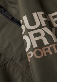 Olivengrønt jakke stof med gittertekstur, der har et stort hvidt og beige "Superdry" logo. Lynlåsdetaile synlig langs den øverste kant.