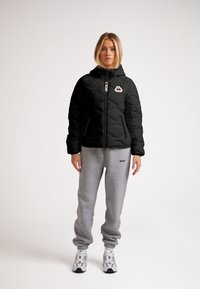 Giacca nera imbottita con cappuccio e polsini a coste, dotata di zip frontale e logo. Abbinate a pantaloni tuta grigi chiari e scarpe da ginnastica bianche.