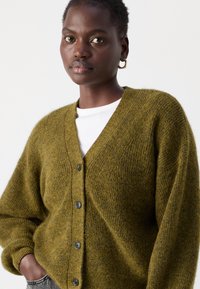 Πράσινο πλεκτό καρντάν με v-λαιμόκοψη, κουμπιά μπροστά και υπερμεγέθεις μανίκια; διαθέτει μαλακή υφή και τέσσερα μαύρα κουμπιά.