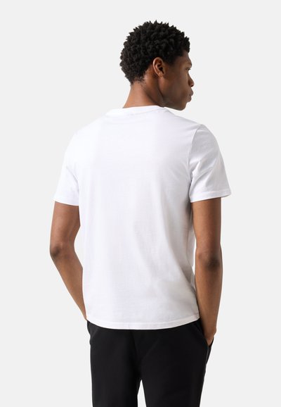Jeune homme aux cheveux courts et bouclés portant un t-shirt blanc uni à manches courtes et un pantalon noir, vu de dos.