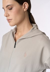 Felpa con zip di colore grigio chiaro realizzata in tessuto morbido, con un logo ricamato discreto sul lato sinistro del petto e un design del cappuccio rilassato.