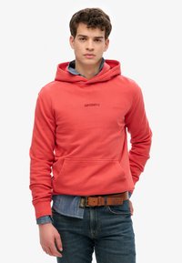 Sudadera de color coral con bolsillo canguro, puños acanalados y cordón ajustable. Llevada sobre una camisa de mezclilla y combinada con jeans oscuros.