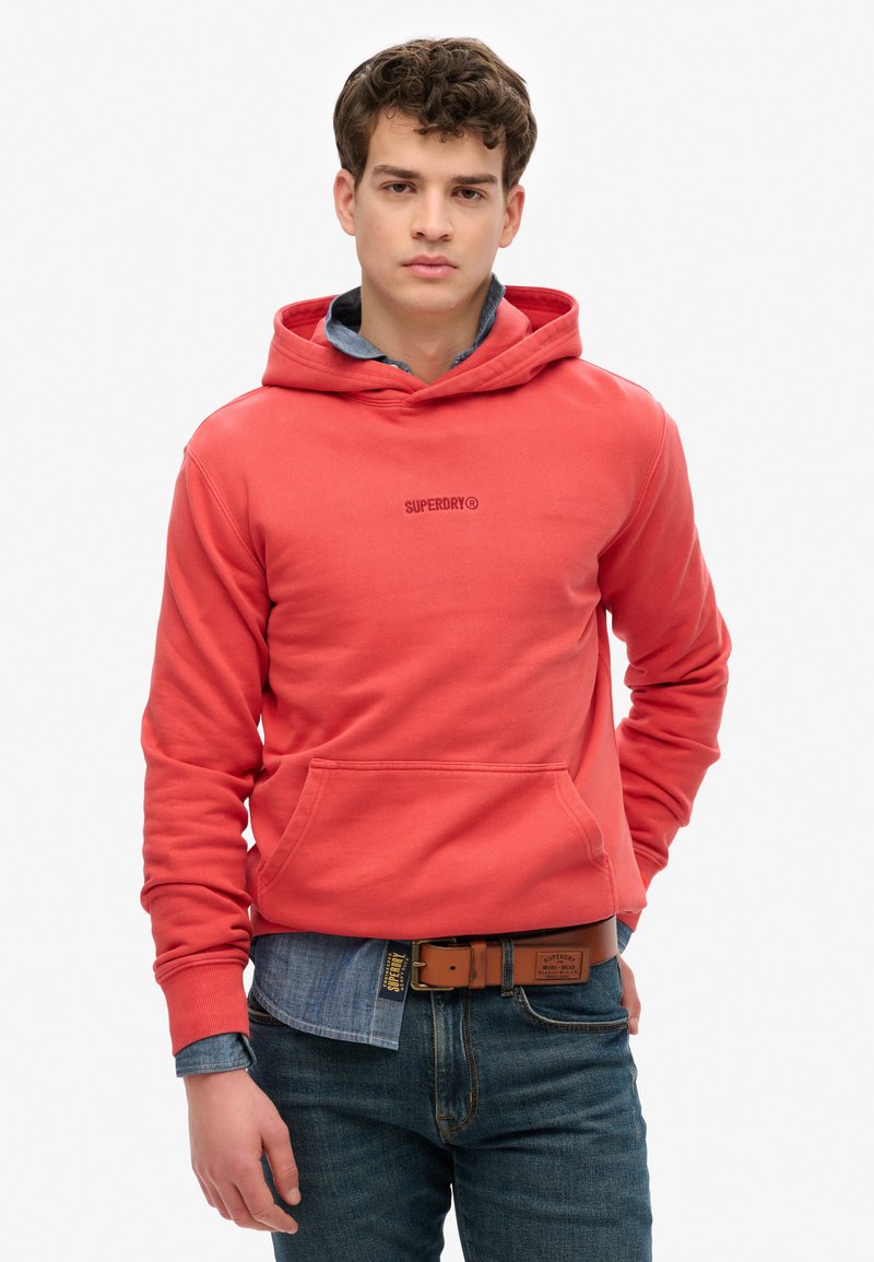 Sudadera de color coral con bolsillo canguro, puños acanalados y cordón ajustable. Llevada sobre una camisa de mezclilla y combinada con jeans oscuros.
