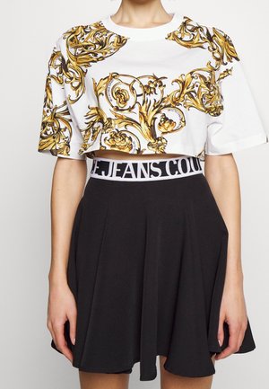 Vrouw draagt een wit cropped shirt met gouden barokpatronen en een zwarte rok met een witte tailleband met het label "JEANS COUTURE."