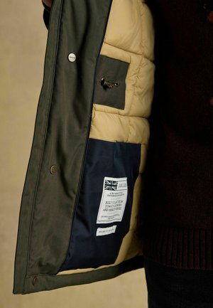 Zelená vonkajšia bunda s béžovým polstrovaným podšívkou a tmavomodrou vnútornou kapsou. Štítok zobrazuje logo značky a pokyny na údržbu.