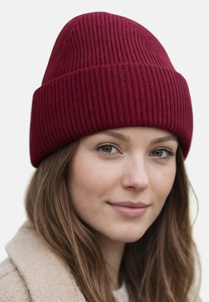 Frau mit langen braunen Haaren, die eine burgunderrote gestrickte Mütze und einen beigen Mantel trägt, lächelt sanft vor einem schlichten hellen Hintergrund.