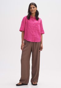 Camicia in lino rosa a maniche corte con colletto e due tasche frontali, abbinata a pantaloni larghi marroni e sandali neri.