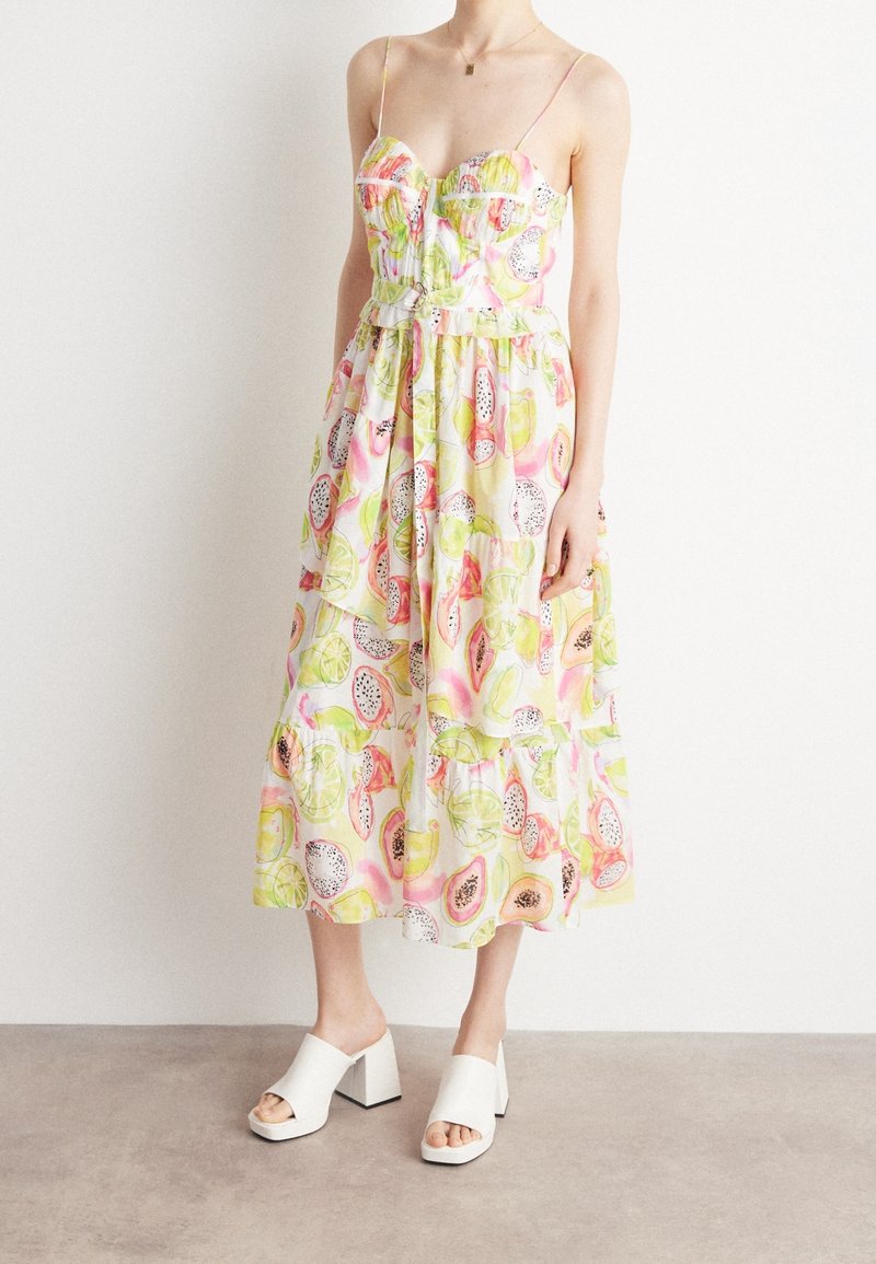Robe midi fleurie avec bretelles spaghetti, présentant un mélange de motifs de fruits colorés sur fond blanc, et une jupe à volants. Mules à talons blocs blanches.