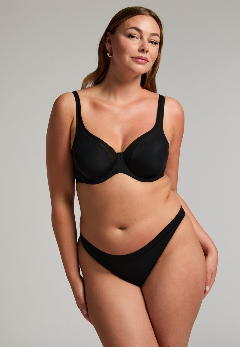 Frau mit langen braunen Haaren, die einen schwarzen Bügel-BH und passende Bikini-Unterwäsche vor einem einfarbigen hellgrauen Hintergrund trägt.