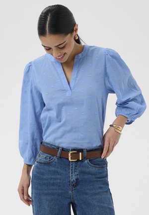 Femme aux cheveux foncés portant un chemisier bleu clair à manches bouffantes, des boucles d'oreilles en or, un bracelet en or, une ceinture marron et un jean bleu, regardant vers le bas.