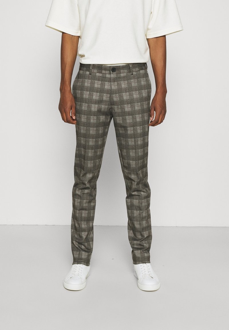 Personne portant un pantalon slim gris à carreaux, des baskets blanches et un top blanc à manches courtes, debout devant un fond uni.