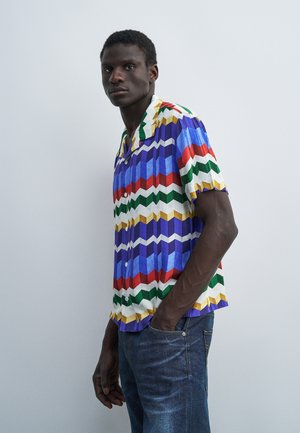 Homme debout contre un mur uni, portant une chemise colorée à manches courtes avec des motifs géométriques en zigzag et un jean bleu.