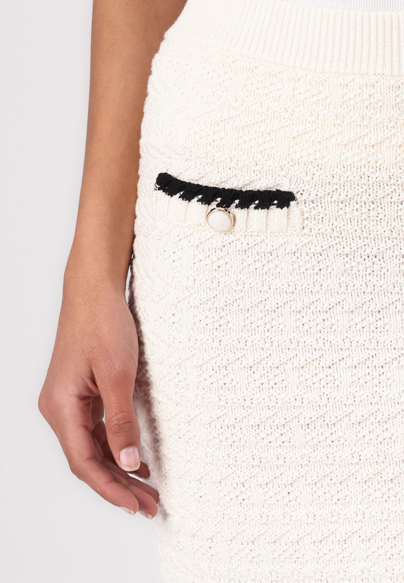 Sweterkowa spódnica w kolorze kremowym z fakturowanym wzorem, czarną kieszenią z okrągłym guzikiem oraz ściągaczem w talii. Ręka umieszczona w pobliżu kieszeni.