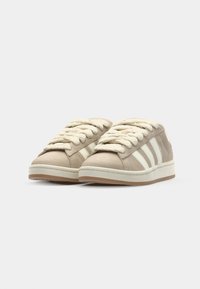 Sneaker beige con una texture liscia simile alla pelle scamosciata, con strisce e lacci color crema, suola in gomma e design minimalista.