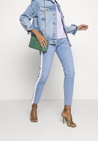 Denimjacke über einem helllila Oberteil, kombiniert mit hellblauen Jeans mit weißen seitlichen Streifen. Eine grüne geflochtene Handtasche und beiger hoher Absatzschuh.
