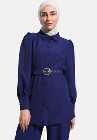 Blusa blu navy con chiusura a bottoni, colletto e maniche a sbuffo. Presenta una cintura con fibbia dorata in vita e una texture di tessuto liscia.