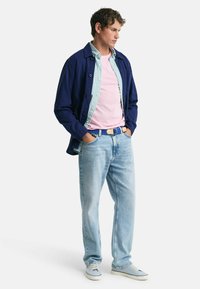 Navy denim jack in gelaagd over een lichtgestreept shirt en een roze t-shirt. Lichtblauwe jeans met een blauwe patronenriem en casual sneakers.