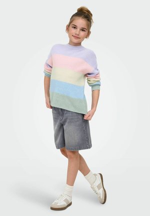 Młoda dziewczyna z włosami upiętymi w kok, ubrana w pastelowy sweter w paski, szare krótkie spodenki do kolan, białe skarpetki i białe trampki, stojąca na jednej nodze.