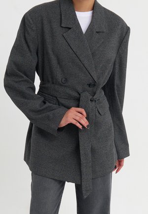 Person wearing a gray checked belted blazer over a white top and gray jeans, with one hand adjusting the belt.