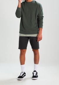 Felpa verde a girocollo indossata sopra una maglietta grigia chiara, abbinata a pantaloni corti scuri e scarpe Vans nere, con calzini bianchi.