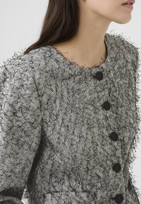 Crno-bijeli kaput u uzorku houndstooth s teksturiranim, resastim rubom i pet crnih dugmadi sprijeda. Prilagođen kroj s okruglim izrezom.