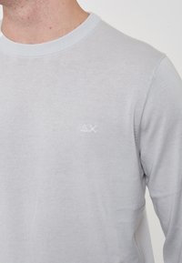 Maglione grigio chiaro a maniche lunghe realizzato in tessuto morbido, con scollo a girocollo e un logo in rilievo discreto sul petto.
