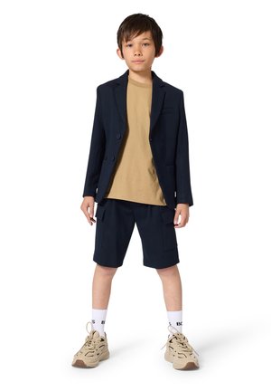 Navy blazer over a tan t-shirt, paired with navy shorts. Sneakers in beige with accents, white socks with black lettering.