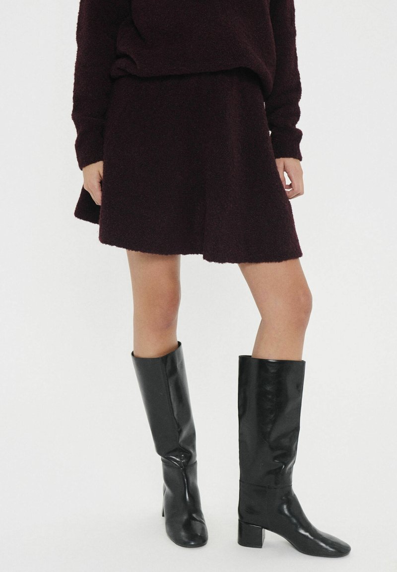 Femme portant un ensemble jupe et pull marron foncé, associé à des bottes en cuir noires montantes jusqu'aux genoux, debout devant un fond blanc.