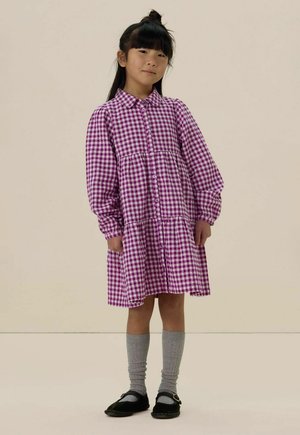 Robe en vichy violet et blanc avec des manches longues, un devant à boutons et des poches, associée à des chaussettes grises montantes et des chaussures noires.