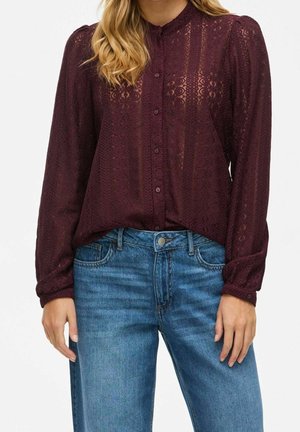 Frau trägt eine langärmelige, burgunderrote Spitzenbluse mit Knöpfen, in blaue Jeans gesteckt, vor weißem Hintergrund.